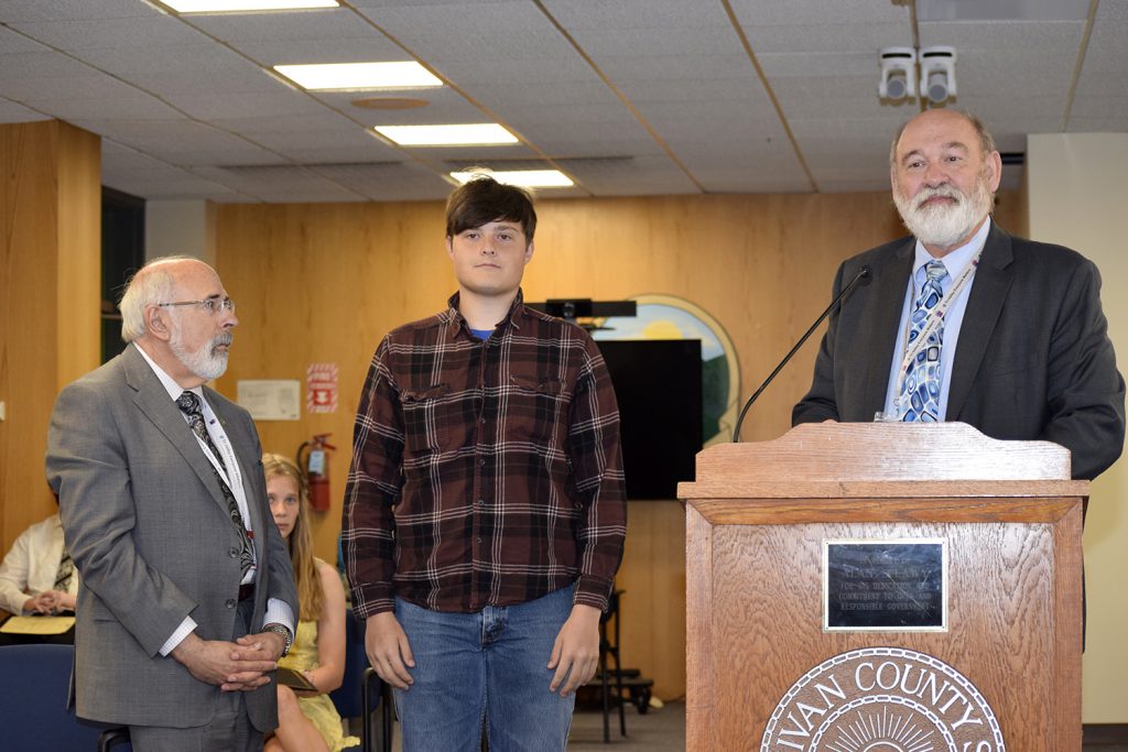 One individual speaking at a podium, two individuals looking on